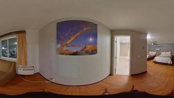 A panoramic view of a hotel room with a large window, a painting of a tree, and a bed with a nightstand.