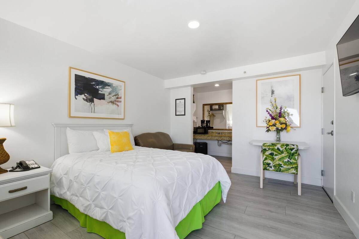 A hotel room with a bed, a nightstand, a chair, a small table with a vase, and a mirror.