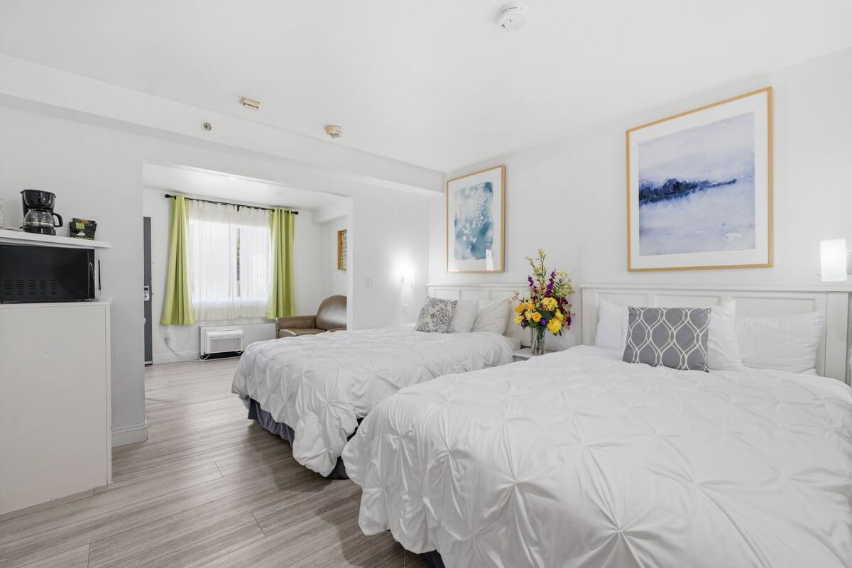 A hotel room with two beds, a kitchenette, a bathroom, and a view of the outdoors.