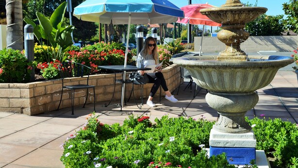 A woman sitting at an outdoor table with a blue and white umbrella, surrounded by a garden with a stone fountain. - Loma Linda Inn (Loma Linda)