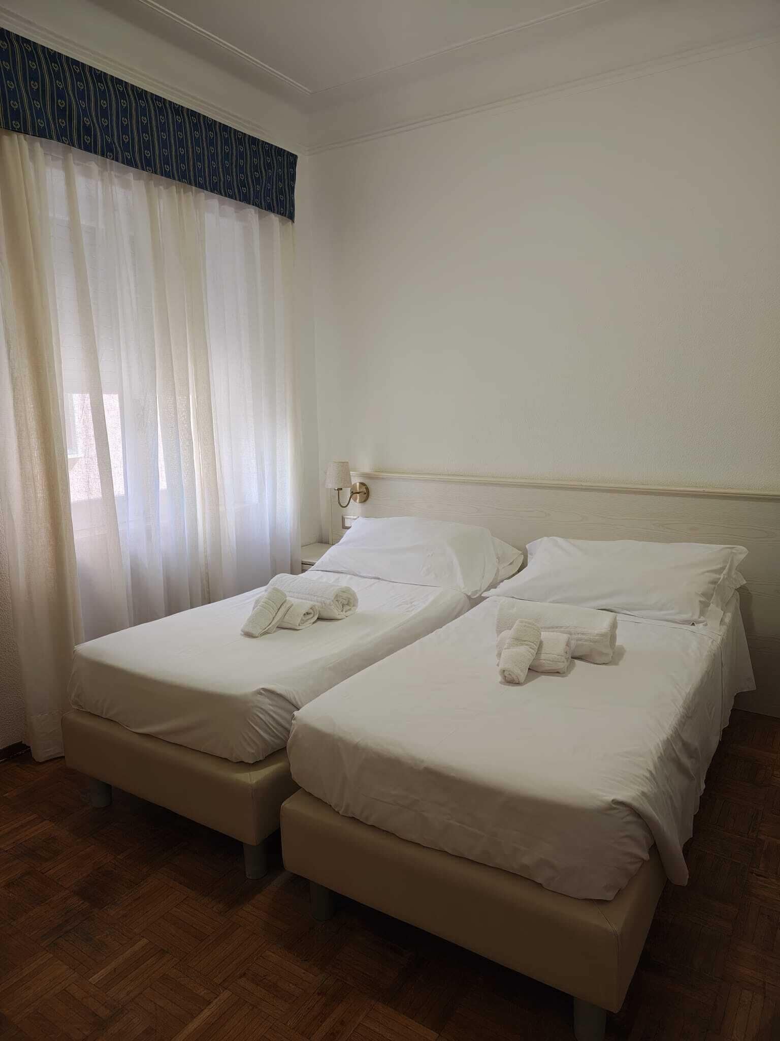 Habitación de hotel con dos camas, piso de madera y ventana con cortinas translúcidas.