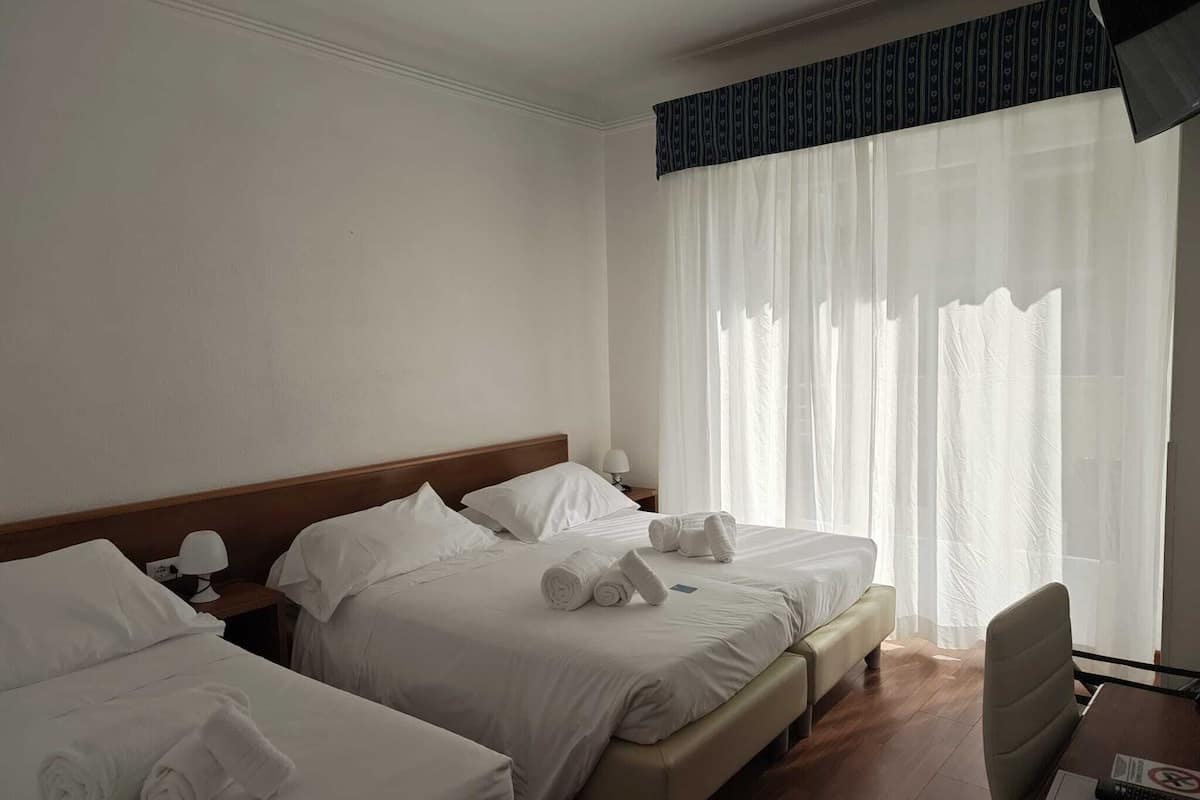 A hotel room with two beds, a wooden headboard, a desk with a chair, and a large window with sheer curtains.