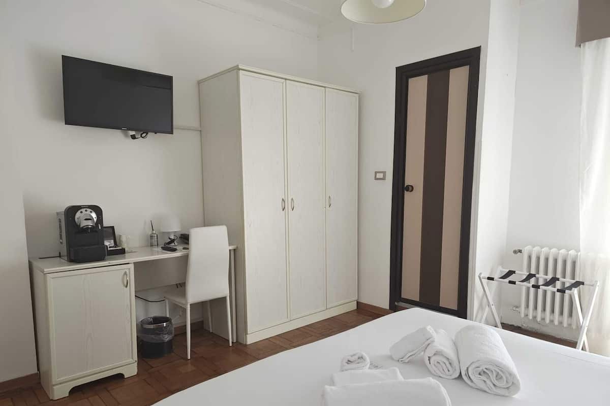 A hotel room with a bed, a desk with a coffee machine, a wardrobe, a television, and a radiator.