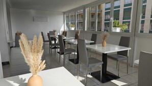A modern dining area with white tables and chairs, large windows, and potted plants. - Hotel Rio (Sanremo)