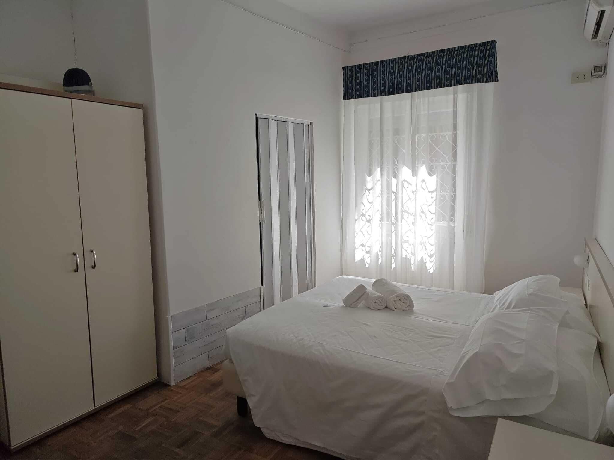 Un dormitorio con cama, armario, ventana con cortinas y radiador.