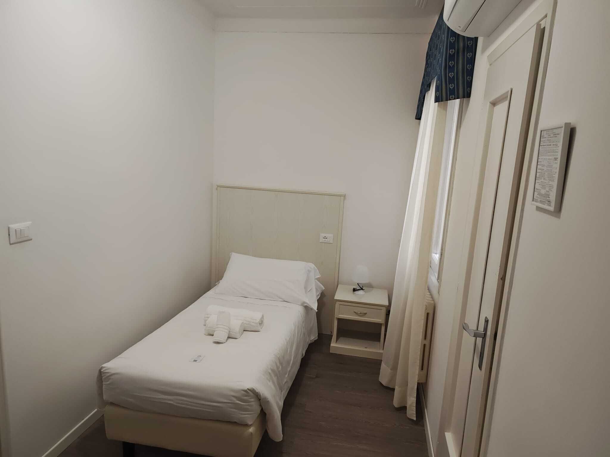 A small, clean room with a single bed, a bedside table, a nightstand, and a door with a curtain.