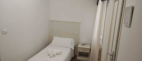 A small, clean room with a single bed, a bedside table, a nightstand, and a door with a curtain.