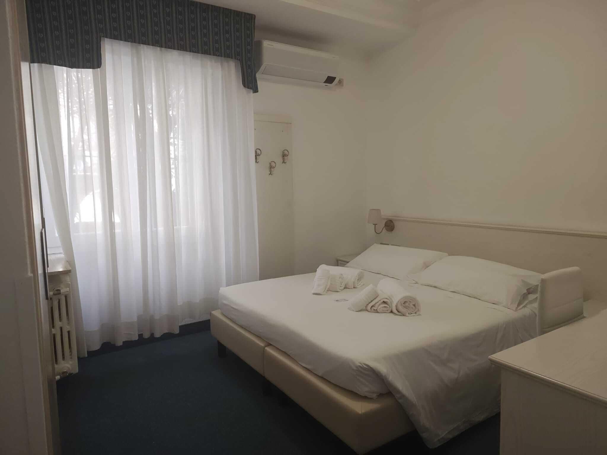 Ein Hotelzimmer mit Bett, Nachttisch, Lampe und Fenster mit durchsichtigen Vorhängen.