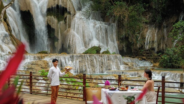 A dining area with a waterfall view, wooden deck, and a table set for two. - La Résidence Phou Vao (Luang Prabang)