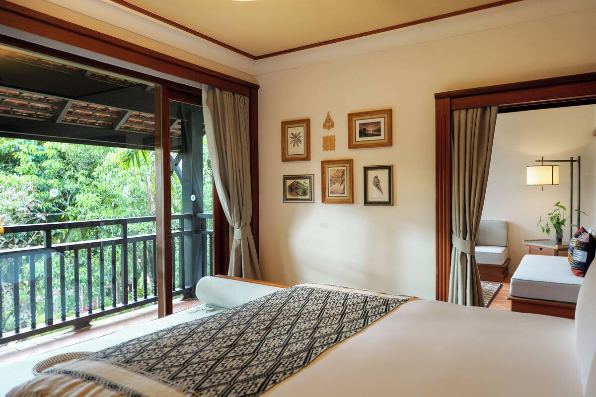 Een hotelkamer met een groot bed, een balkon met uitzicht op groen en verschillende ingelijste foto’s aan de muur.