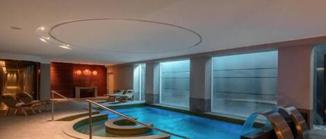 A modern indoor pool area with a central pool, a seating area with lounge chairs, and a staircase leading to the pool.