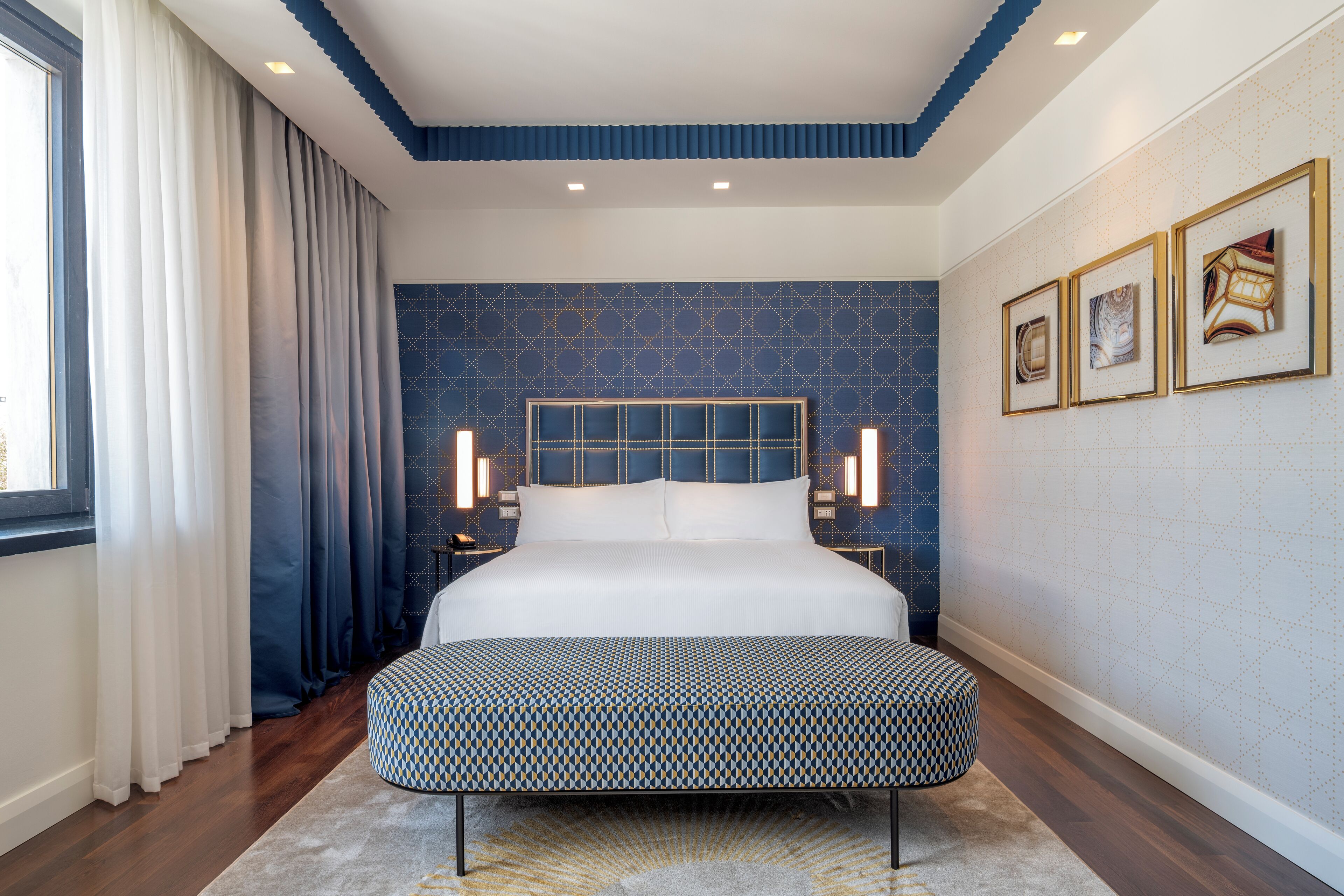 A modern bedroom with a large bed, a bench, a patterned wall, and framed artwork.