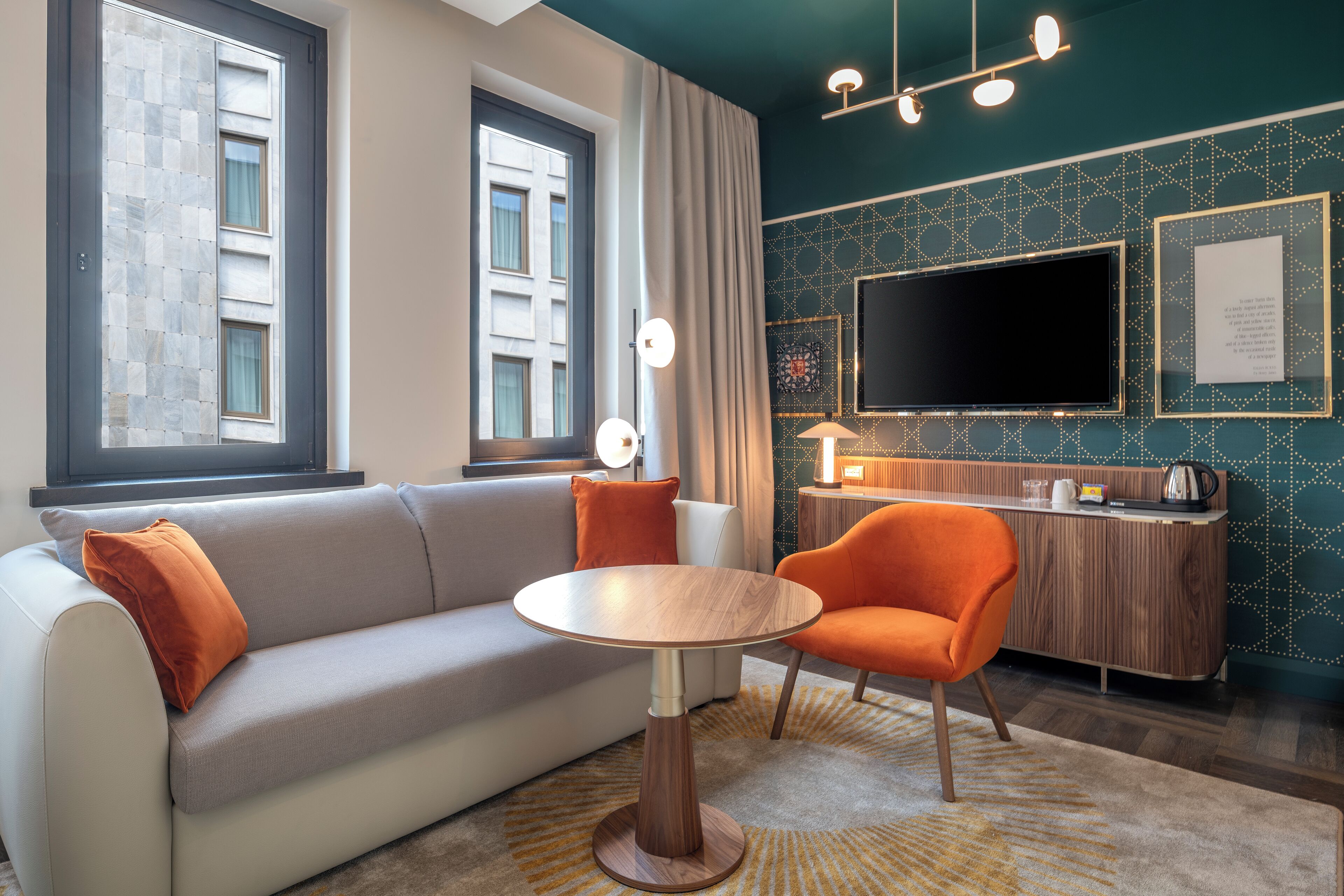 A modern living room with a sofa, a round table, and an orange chair. There is a TV mounted on the wall and a wooden cabinet with a kettle on top.