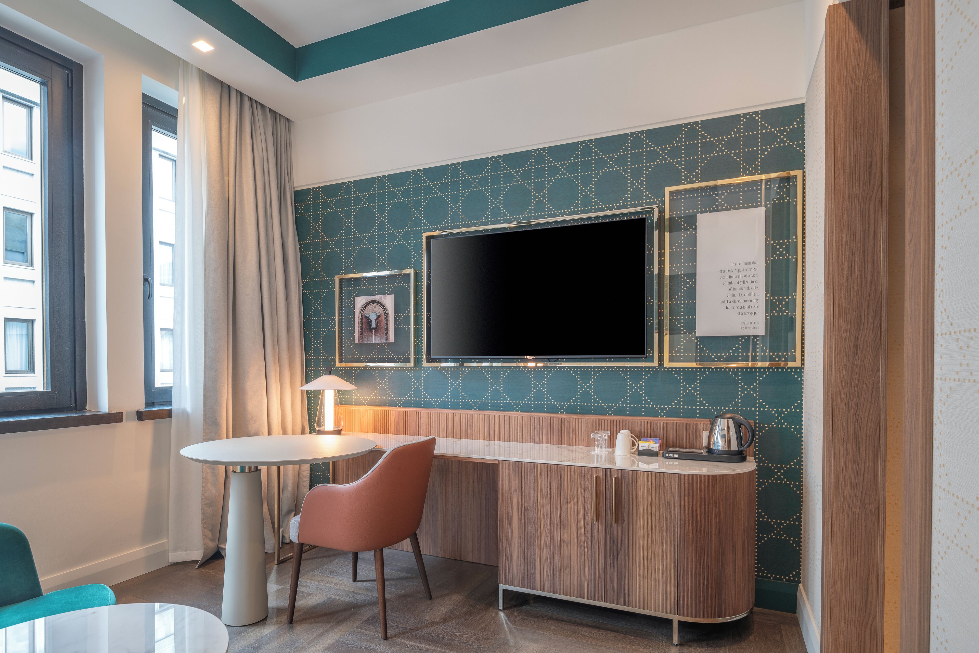 Habitación de hotel con una televisión de pantalla plana grande montada en la pared, una mesa redonda con una silla, un aparador de madera con un hervidor y un cuadro enmarcado en la pared.