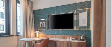Habitación de hotel con una televisión de pantalla plana grande montada en la pared, una mesa redonda con una silla, un aparador de madera con un hervidor y un cuadro enmarcado en la pared.