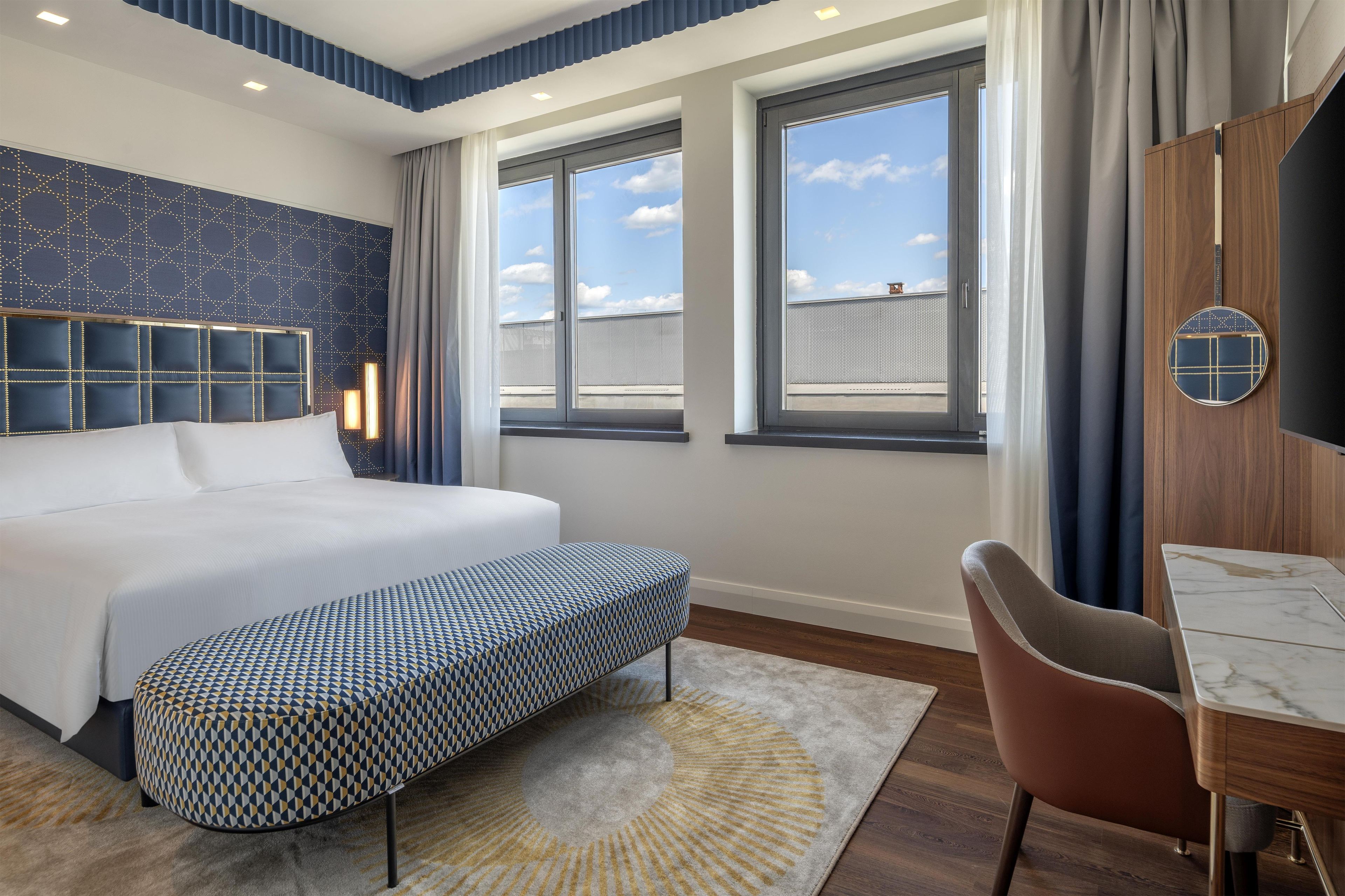 A modern hotel room with a large bed, a seating area, and a view of the sky through the windows.