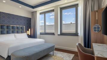 A modern hotel room with a large bed, a seating area, and a view of the sky through the windows.
