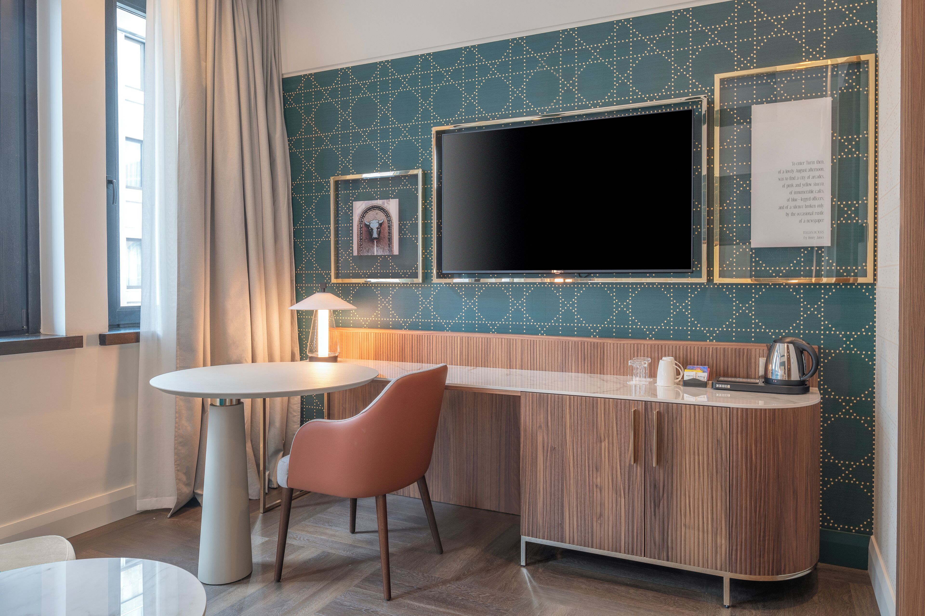 Quarto de hotel moderno com uma mesa redonda, uma poltrona acolchoada, um armário de madeira, uma TV de tela plana e uma parede decorativa com padrão geométrico.