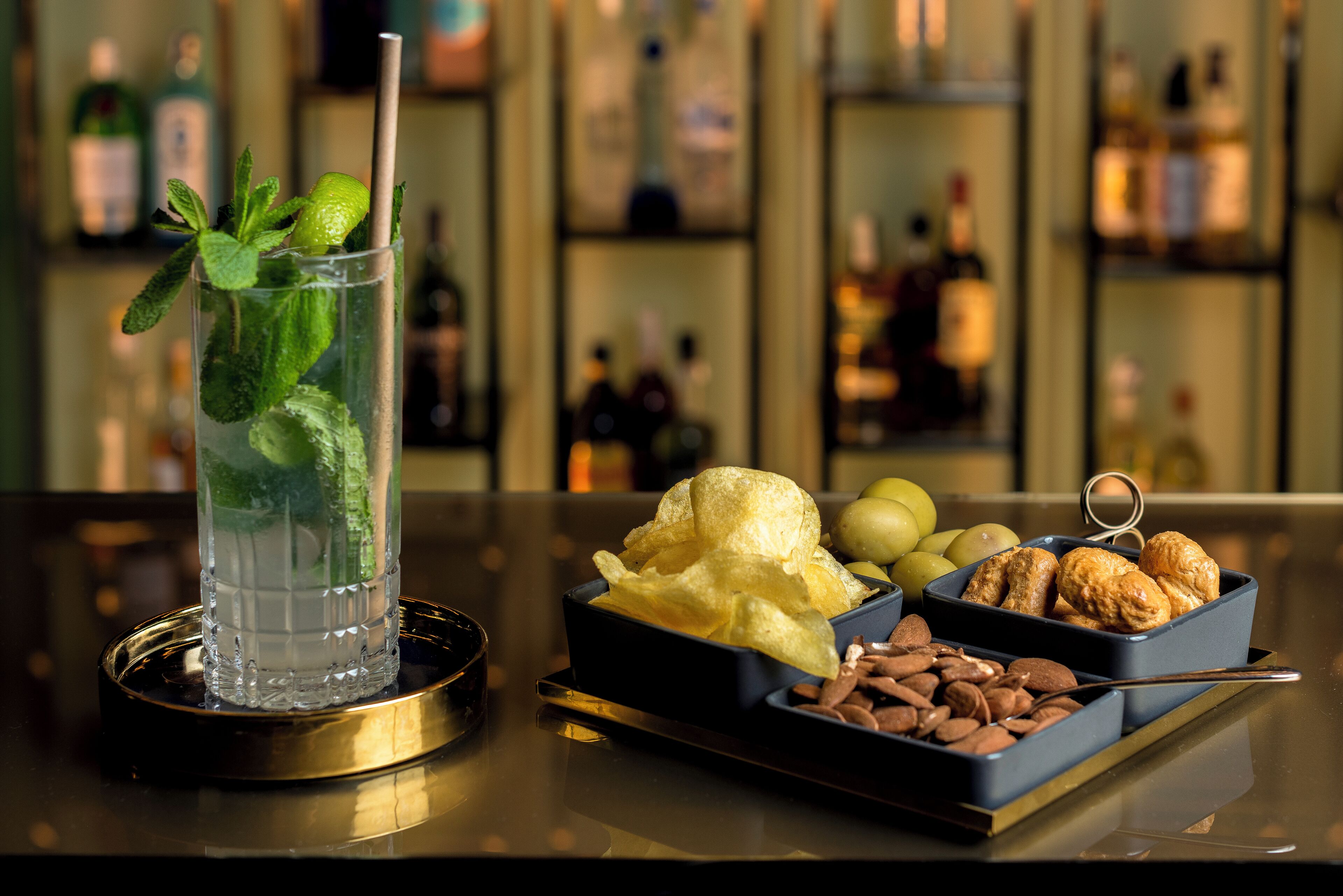 A bar setting with a glass of a clear beverage garnished with mint, a bowl of chips, and a tray of assorted nuts and chocolates.