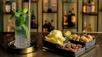 A bar setting with a glass of a clear beverage garnished with mint, a bowl of chips, and a tray of assorted nuts and chocolates.