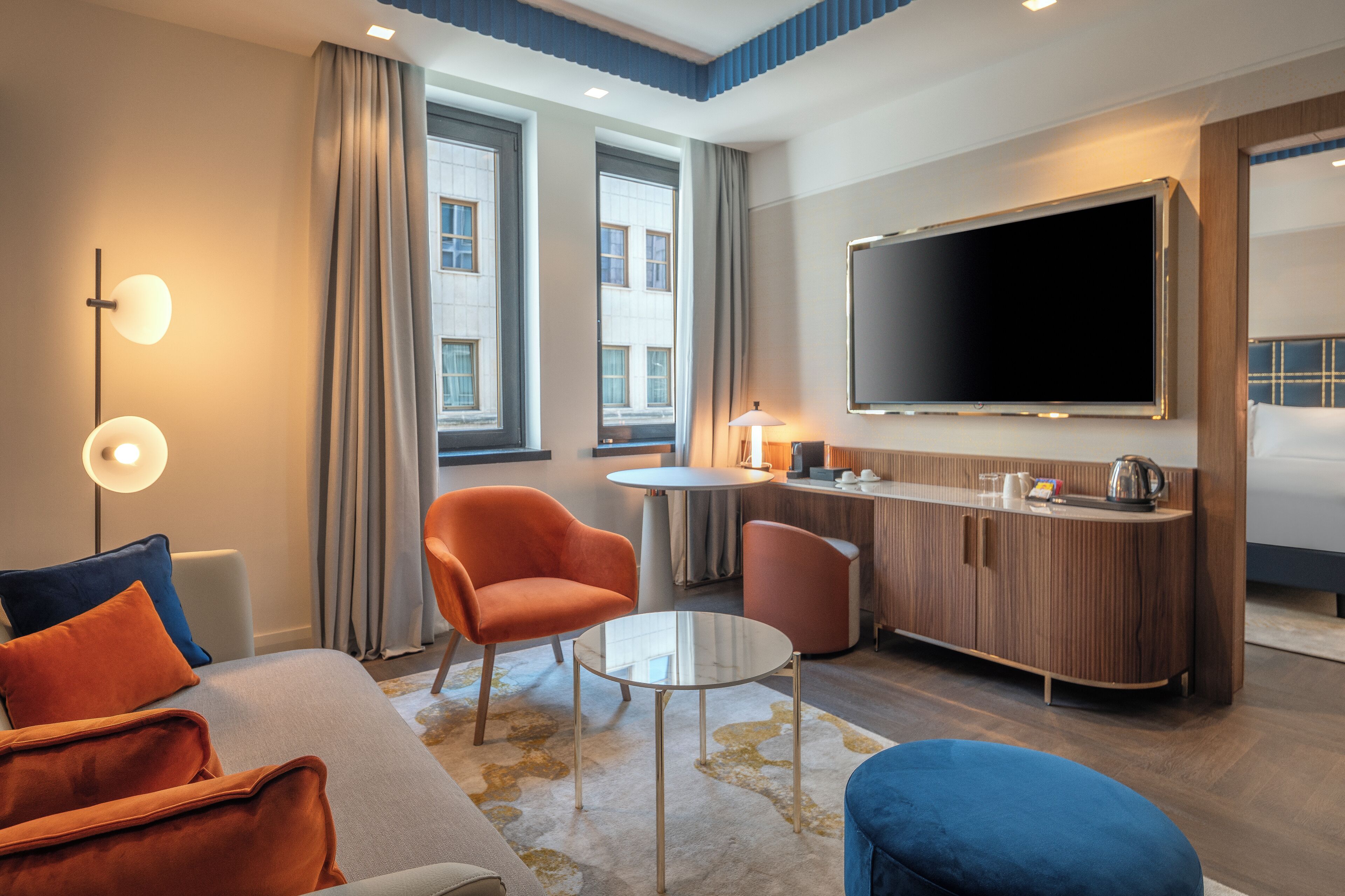 Ein modernes Hotelzimmer mit einer Sitzecke, zwei Sesseln, einem runden Couchtisch, einem Fernseher und einer Minibar.