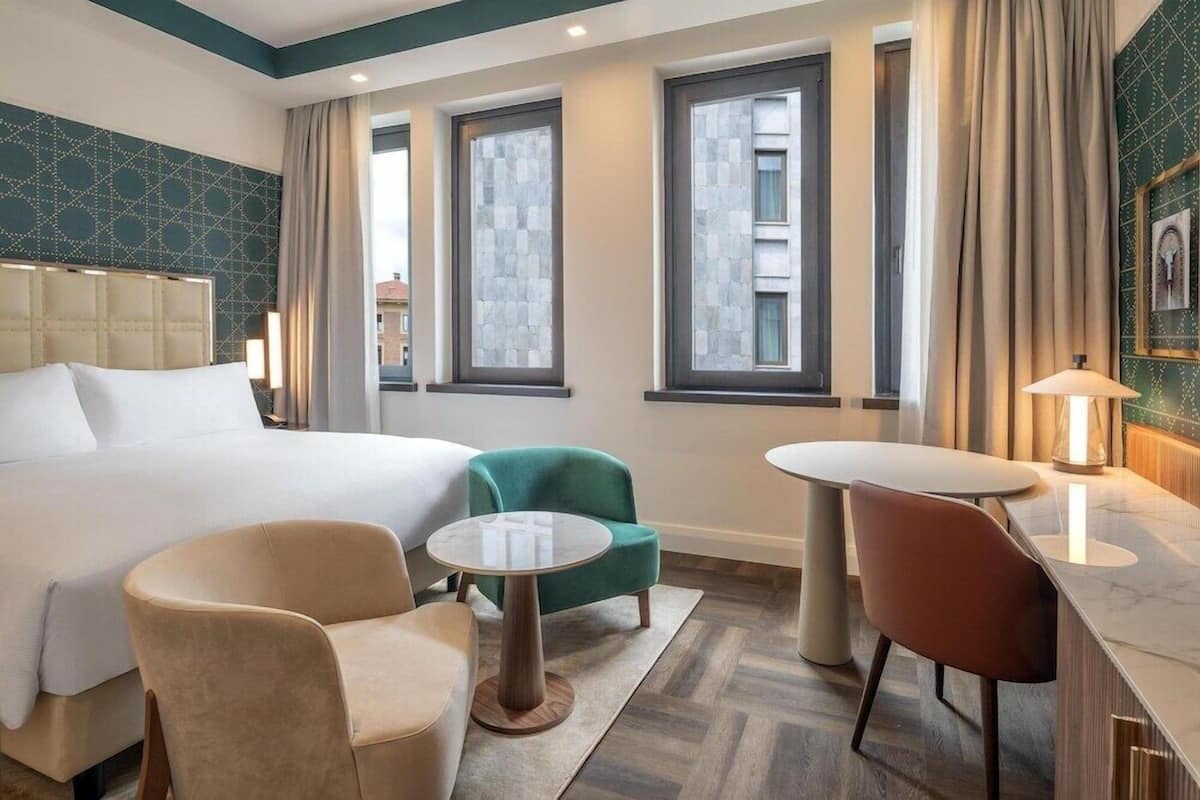 A hotel room with a bed, two chairs, a small table, and three windows.