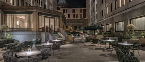 Un espacio al aire libre con mesas y sillas redondas, rodeado de edificios con grandes ventanales.
