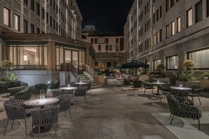 An outdoor seating area with round tables and chairs, surrounded by buildings with large windows. - Hilton Turin Centre (Turin)
