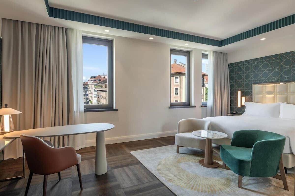 A hotel room with a bed, two chairs, a small table, and a view of buildings through large windows.