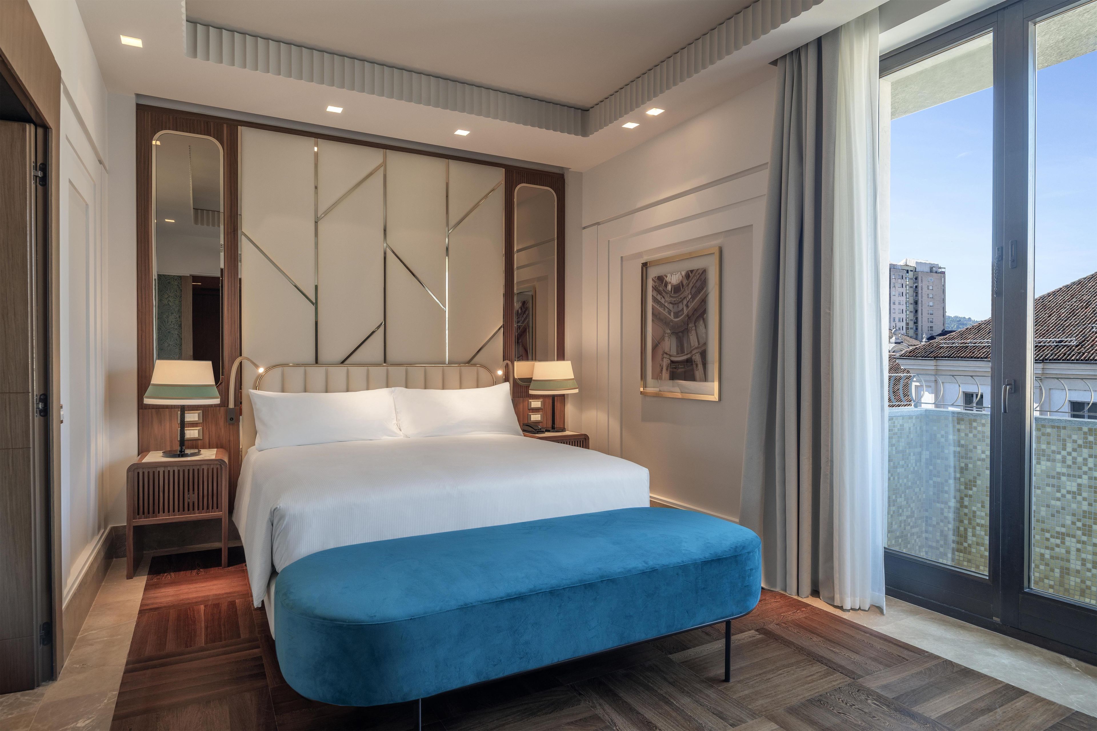 A modern hotel room with a large bed, a blue bench, a city view, and a large mirror.