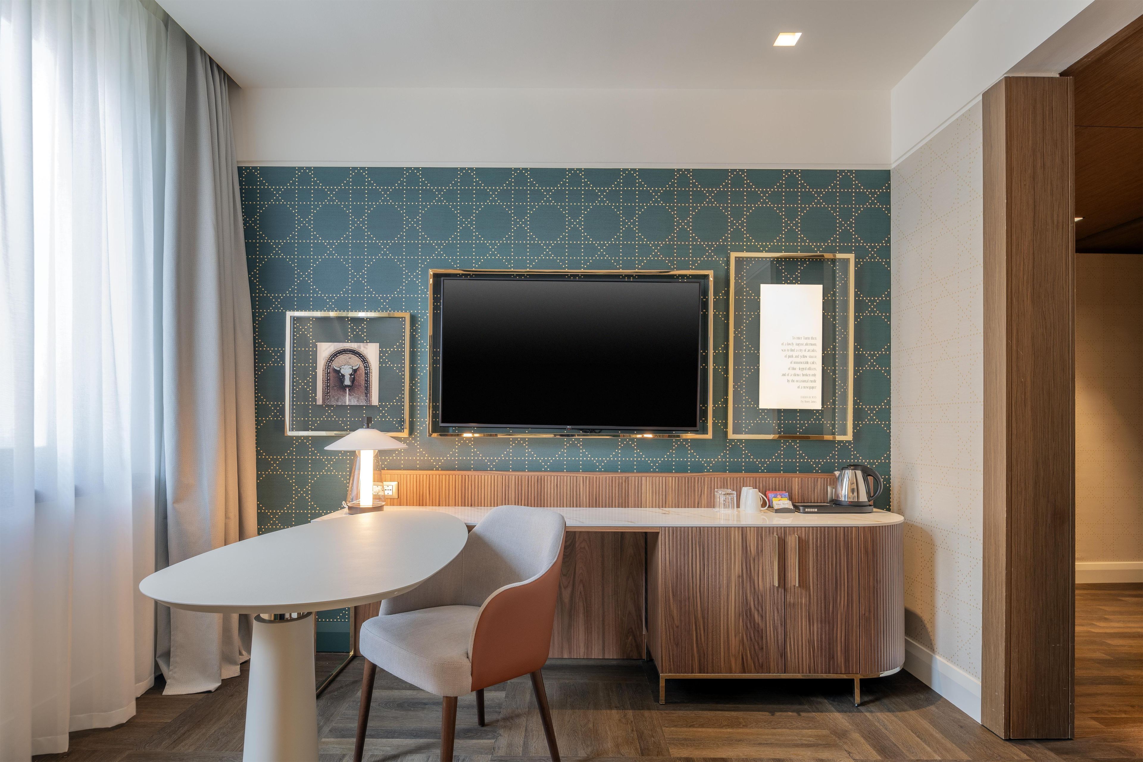 Habitación de hotel con televisor de pantalla plana montado en la pared, escritorio de madera con una silla, mesita con lámpara y un cuadro enmarcado en la pared.