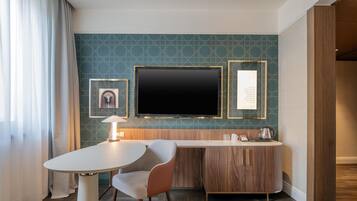 Habitación de hotel con televisor de pantalla plana montado en la pared, escritorio de madera con una silla, mesita con lámpara y un cuadro enmarcado en la pared.
