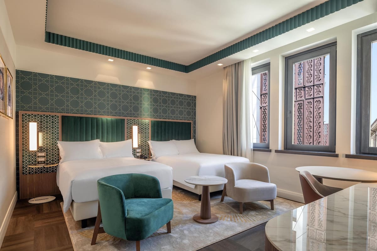 A hotel room with two beds, a green armchair, a round table, and a view of the outside through large windows.