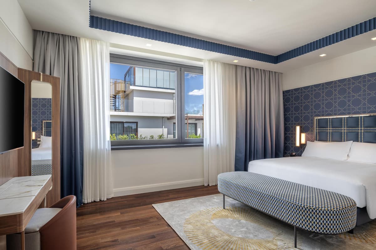 A modern hotel room with a large bed, a desk, and a view of the cityscape.