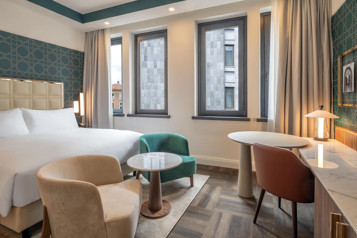 A hotel room with a large bed, two armchairs, a small round table, and a dining table. There are three windows with curtains, a patterned wall, and a framed picture on the wall.
