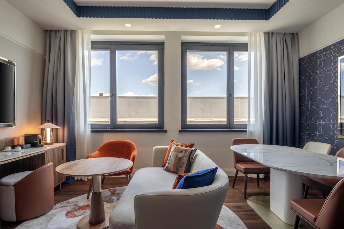 A modern hotel room with a large window, a round dining table, a sofa, and a TV on the left wall.