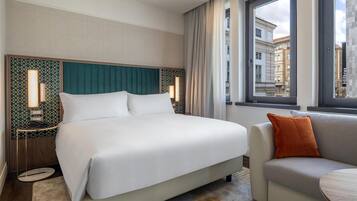 A hotel room with a large bed, a sofa, a nightstand with a lamp, and a view of buildings through the window.