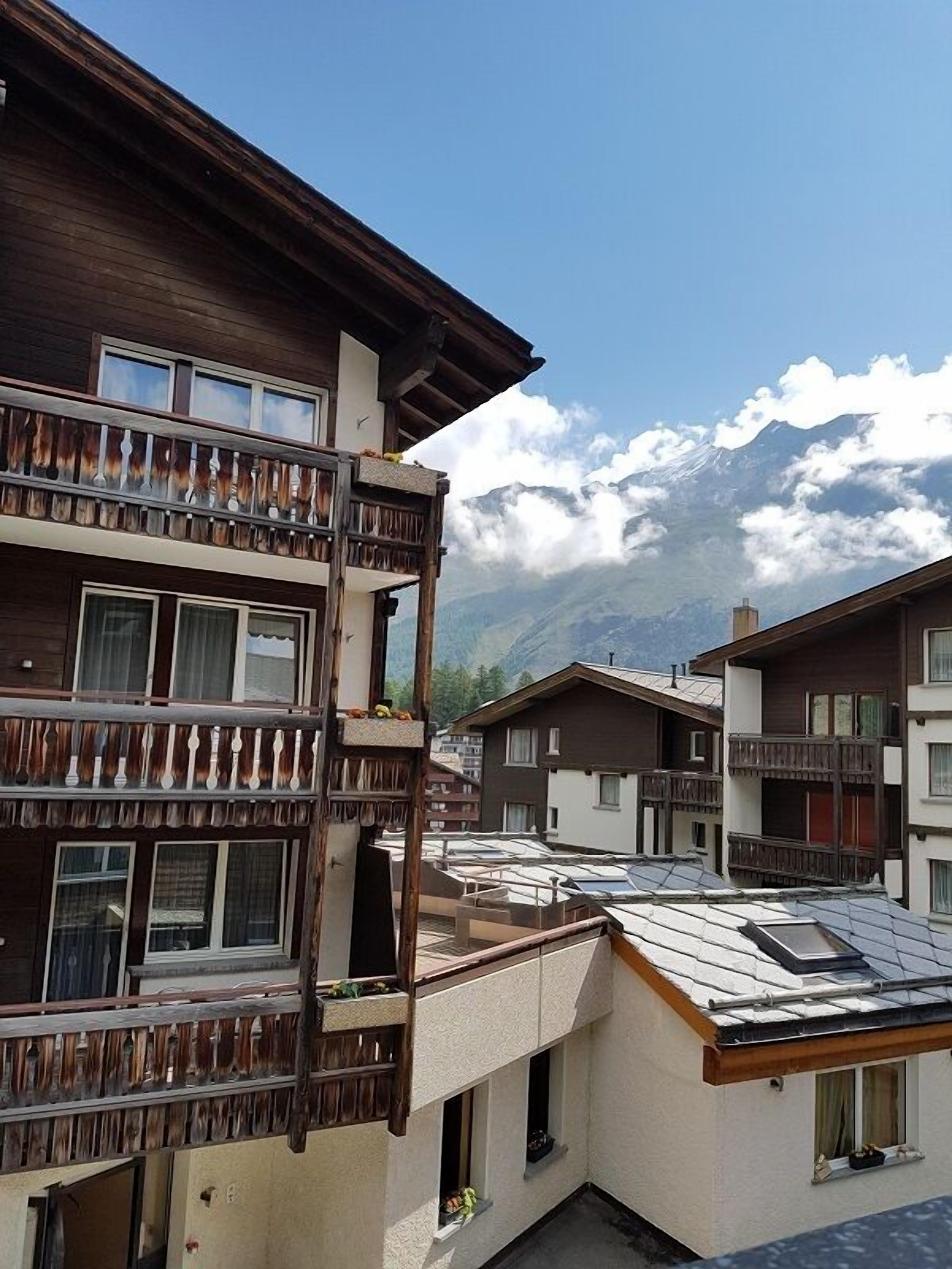 A residential building with balconies, a sloped roof, and a mountainous backdrop.