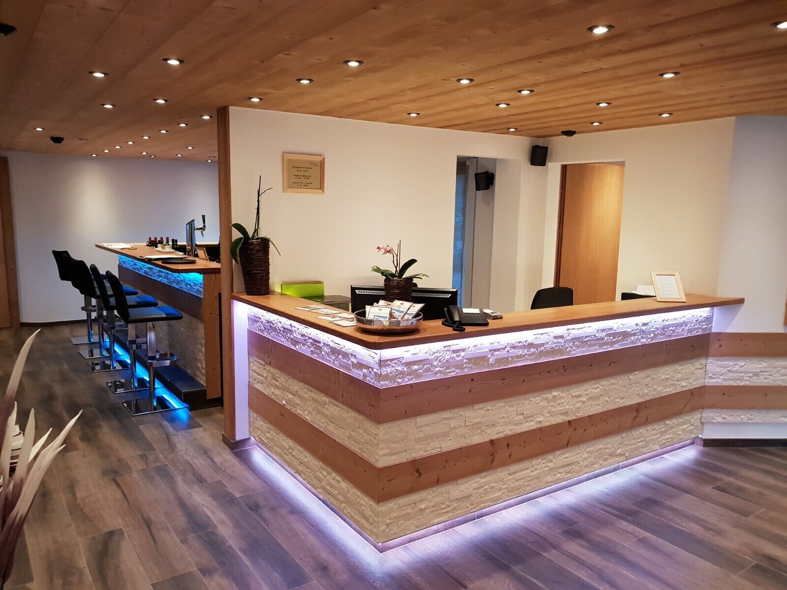 A reception desk with a layered design, a bar area with stools, and a wooden ceiling with recessed lighting.