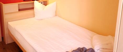 A single bed with white bedding, a bedside table with a lamp, a flat-screen TV mounted on the wall, and a framed artwork above the bed.