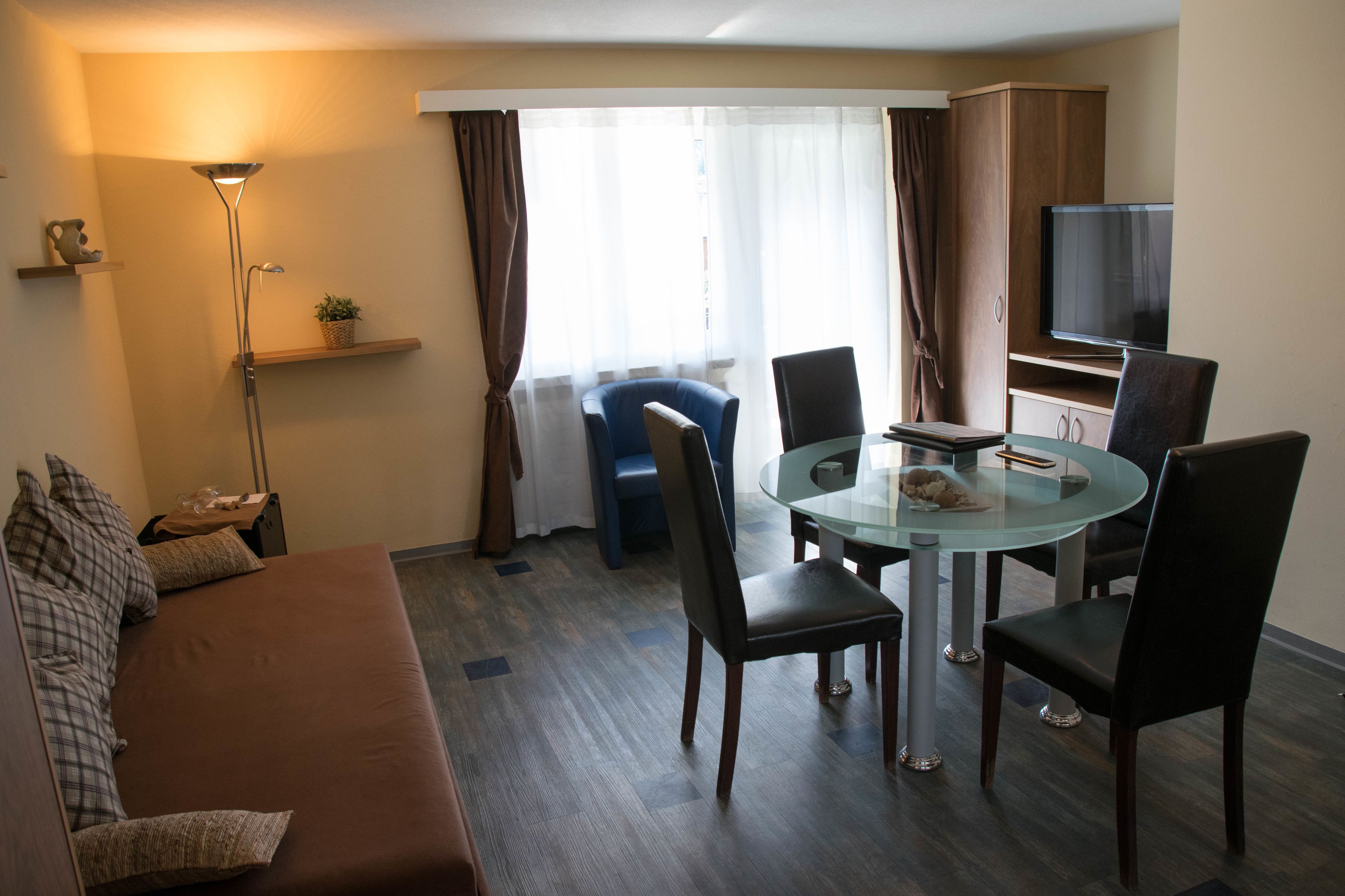 A modern hotel room with a brown sofa, a glass dining table with four chairs, a flat-screen TV, and a large window with curtains.