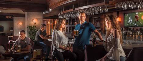 Eine Bar mit verschiedenen Sitzgelegenheiten, einem Fernsehbildschirm, der ein Sportspiel zeigt, und einer Auswahl an Flaschen und Gläsern in den Regalen.