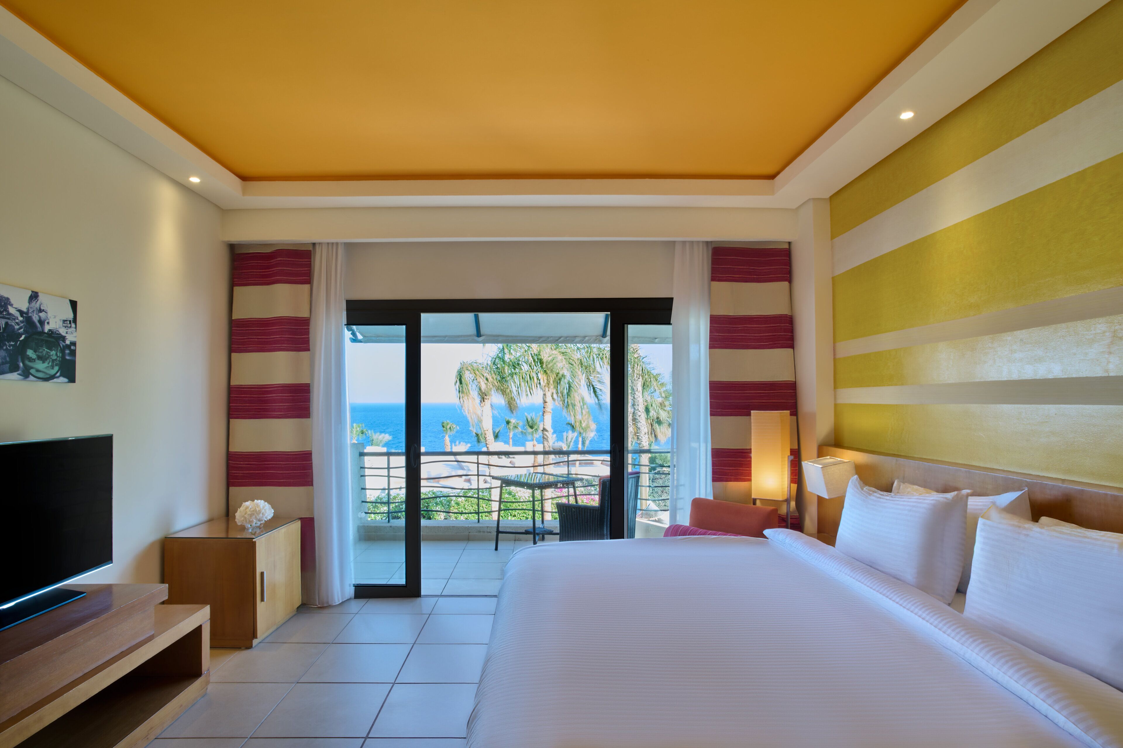 Habitación de hotel con una cama grande, televisión, balcón y vistas al océano, además de cortinas a rayas.