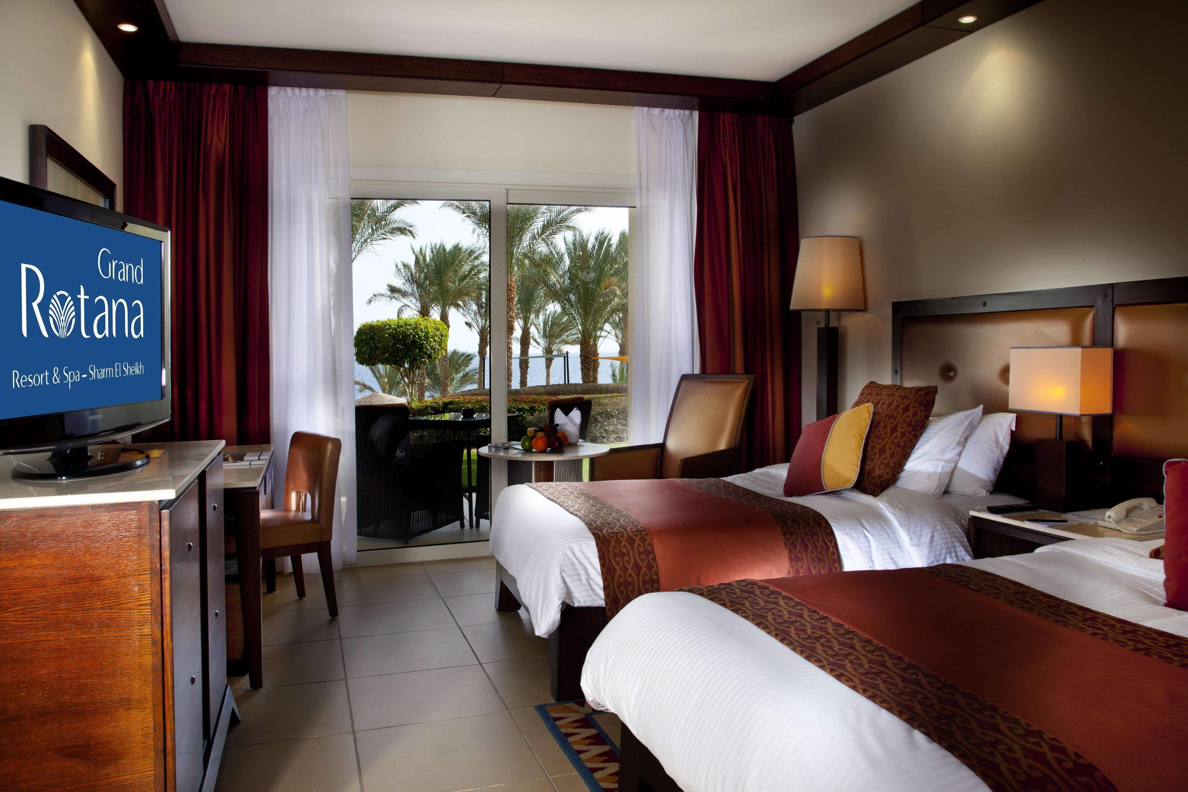 A hotel room with two beds, a desk with a television, a chair, and a view of palm trees and a body of water.