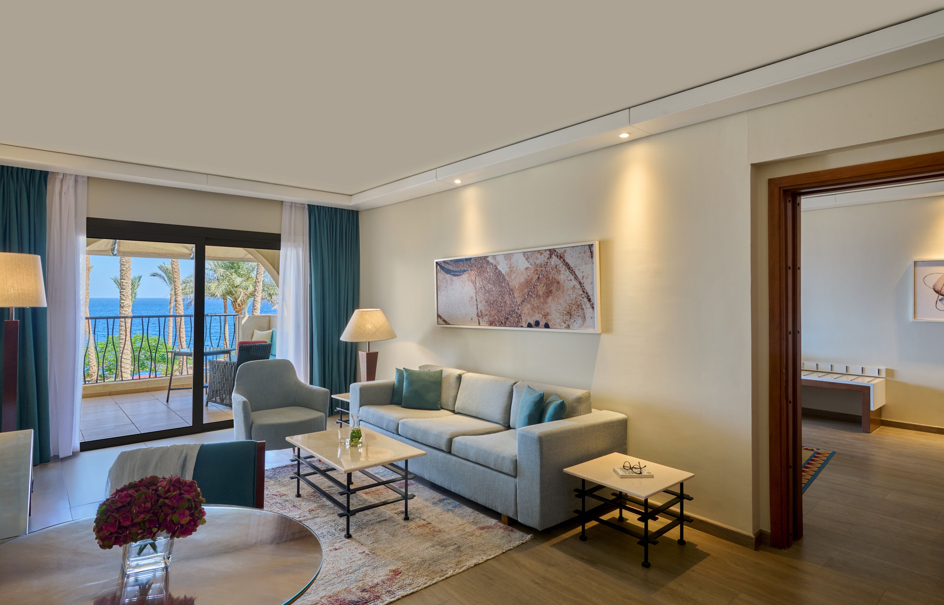 A modern hotel room with a balcony, a sofa, a coffee table, and a painting on the wall.