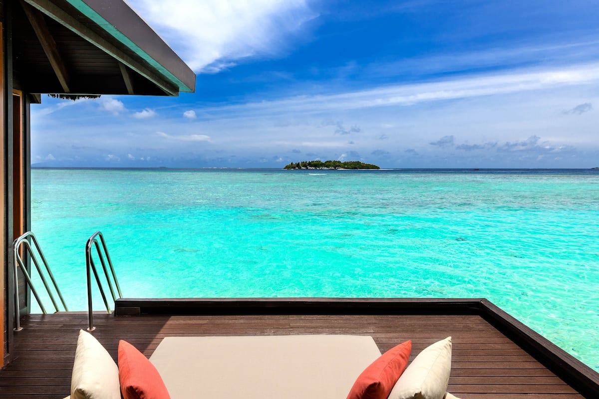 An infinity pool with a wooden deck, white and red cushions, and a view of a turquoise sea and an island.
