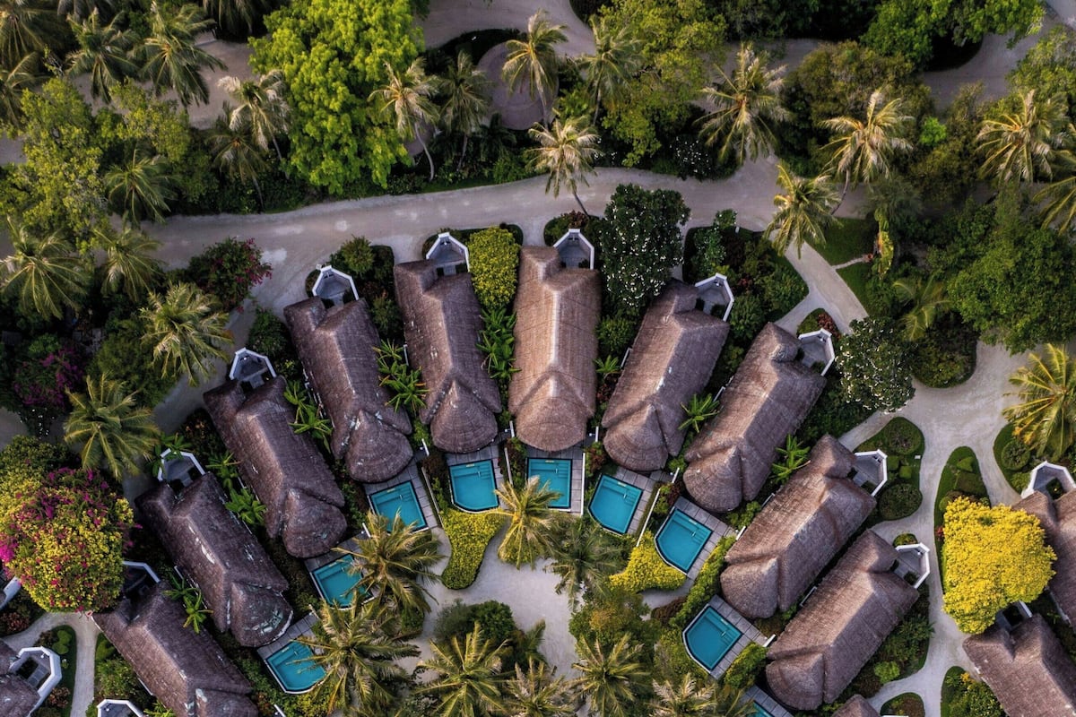 An aerial view of a resort with circularly arranged villas, each with a private pool, surrounded by lush greenery and a sandy beach.