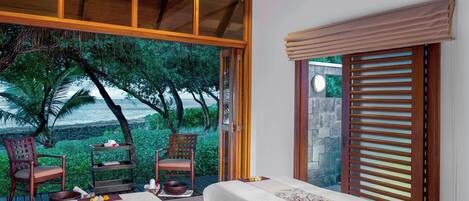 A bedroom with two beds, a wooden ceiling, a large window with a view of trees and the ocean, and a sliding door with blinds.