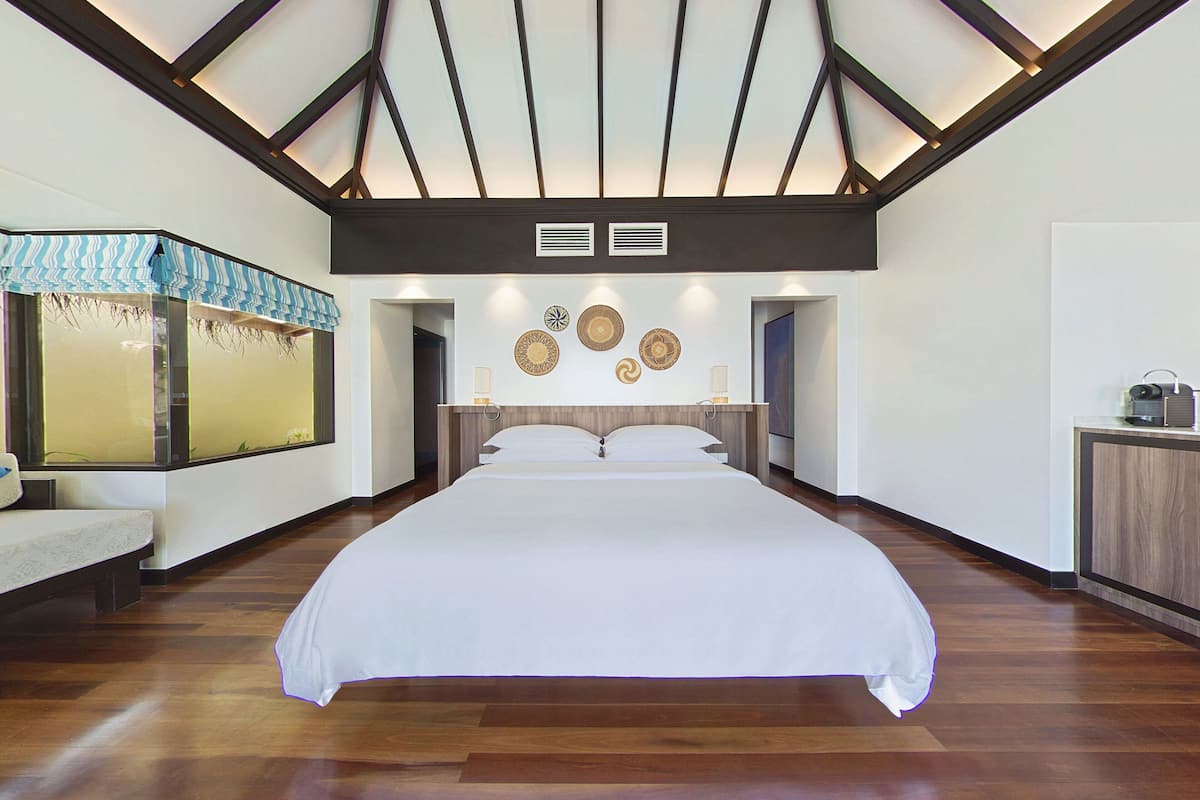 A spacious bedroom with a large bed, wooden flooring, a sloped ceiling with exposed beams, and a large window with a view.