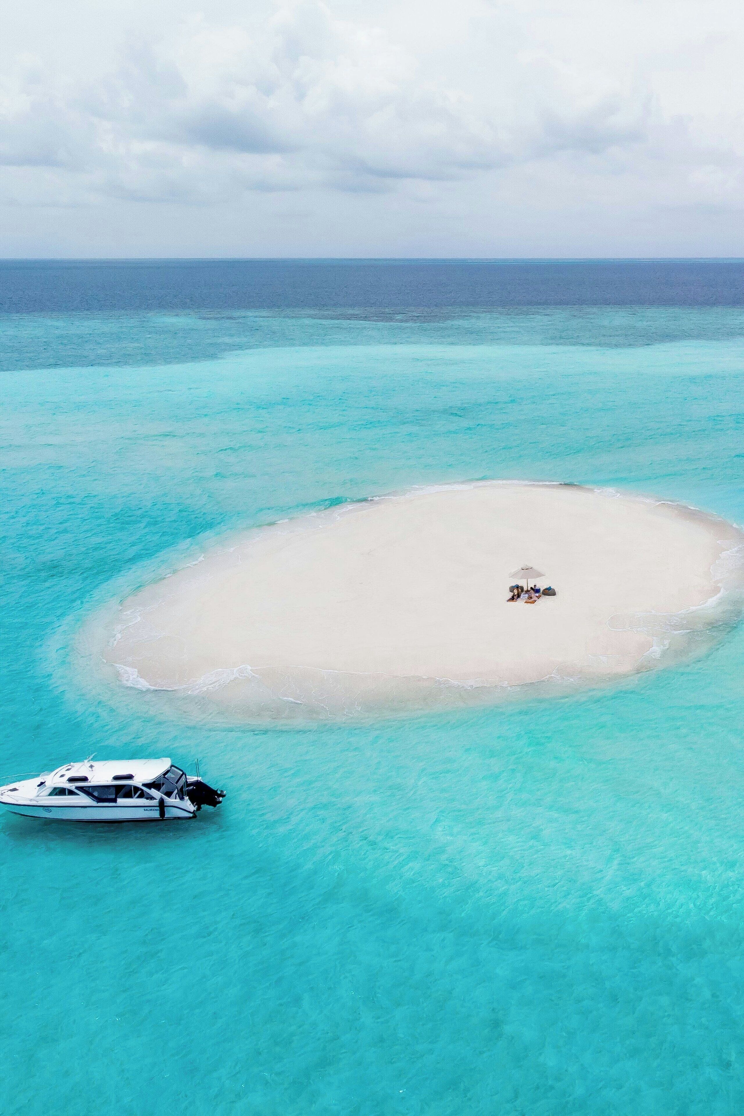 An aerial view of a small, circular island with a sandy beach and a boat anchored nearby.